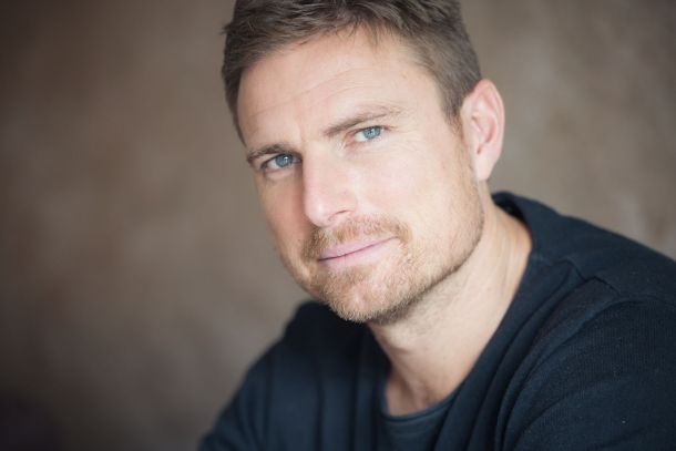 Ein Porträt von Leo Reisinger, dem Autor des Buches *Bavarese*, der mit ernstem Blick und konzentrierter Miene in die Kamera schaut. Er trägt ein dunkles Oberteil. Das Bild bezieht sich auf eine Lesung, bei der er spannende Erzählkunst mit rockiger Live-Musik und Comedy verbindet.